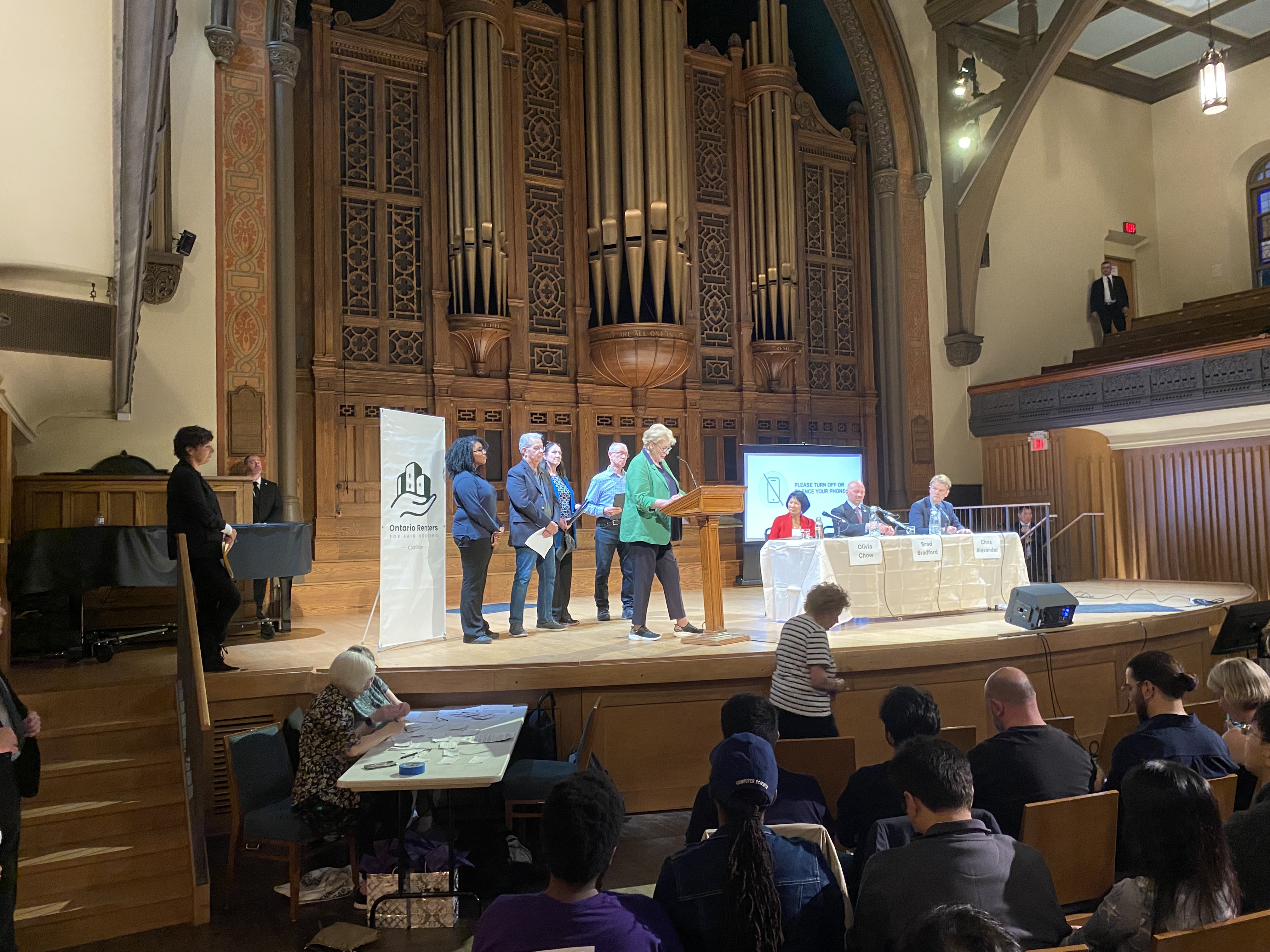 Elizabeth Vonsymborski speaks at the Toronto mayoral debate beside the Ontario Renters Coalition banner, with the candidates on stage.