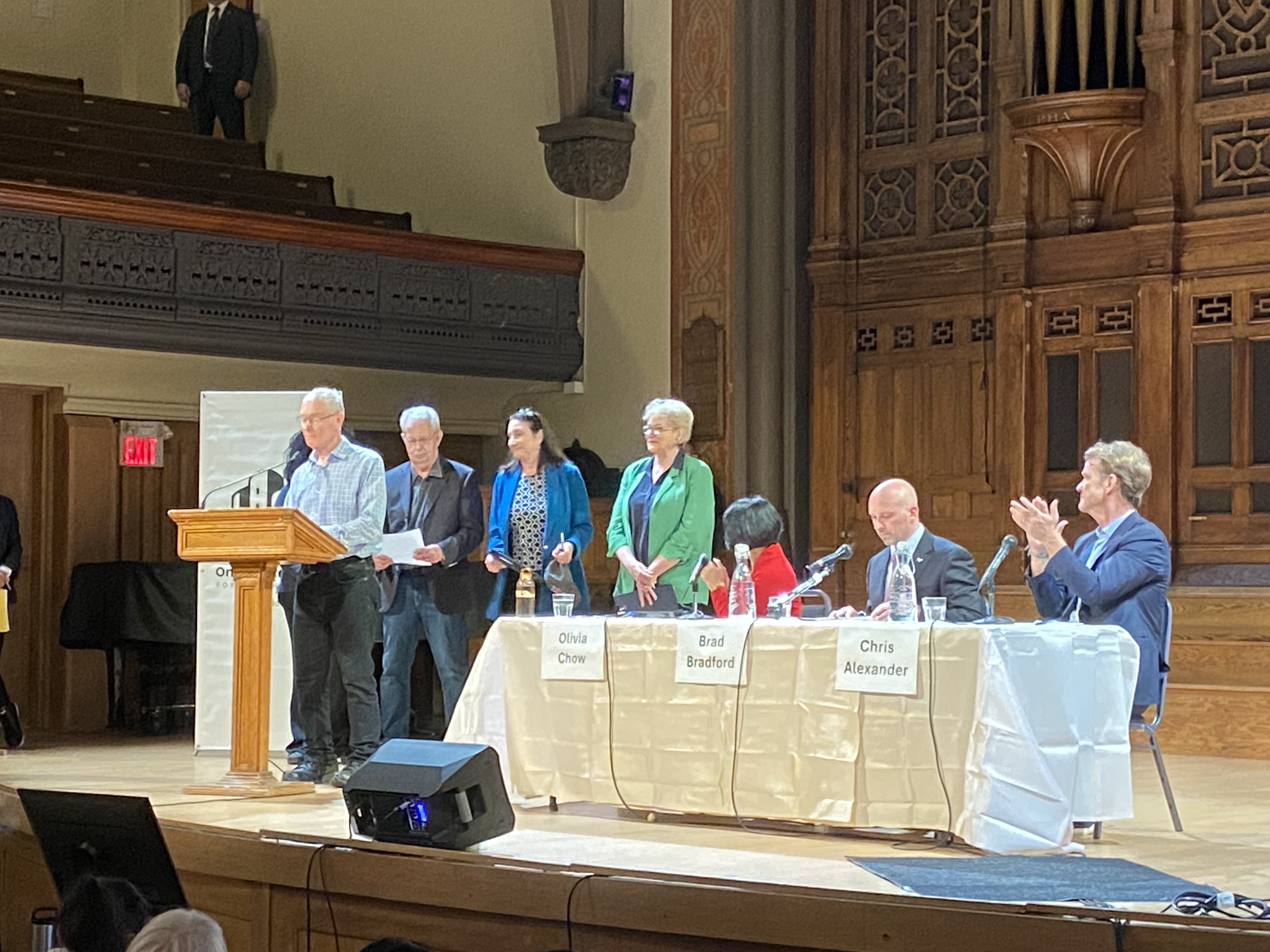 Toronto mayoral candidates seated behind name cards reading Olivia Chow, Brad Bradford and Chris Alexander, with event organizers beside the lectern.